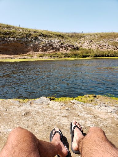 A vertical shot of a male feet sitting on shore of a lake with a hill on the other side