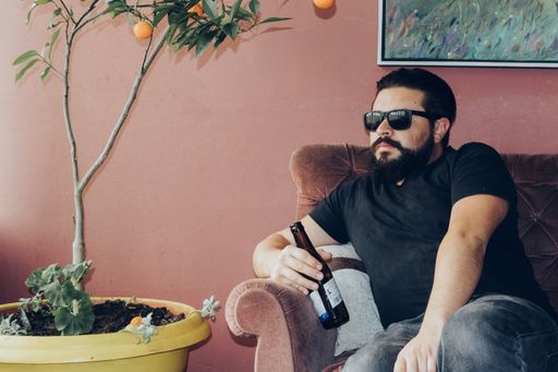 A bearded man wearing shades sitting on the couch while holding his bottled beer
