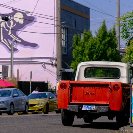 A vibrant pink mural is on display in an urban area, with parked cars and a clear, open skyline in the background