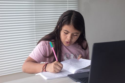 A Hispanic schoolgirl writing in her notebook and doing homework at home with a laptop, the New normal