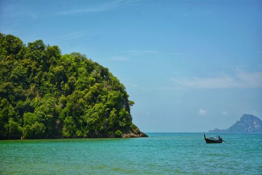 A small boat sailing across the sea with lush trees on the shoreline in Krabi, Thailand.