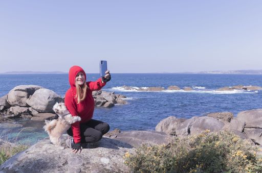 A european female in a red hoodie sitting on a stone by the coast and taking a selfie with her dog