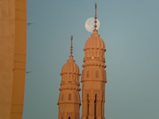 A closeup shot of two towers of different height and the full moon at day time