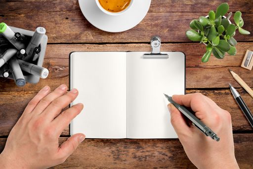 A top view of a male hand writing with a marker on a blank notebook page on wooden desk with pencils, a cup of coffee