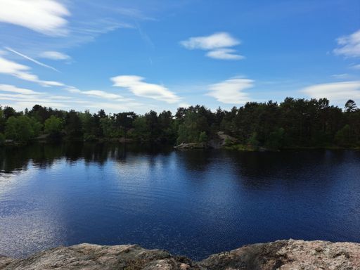 A tranquil lake surrounded by lush greenery, set against a bright blue sky near Kristiansand, Norway.