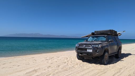 A closeup of a Toyota car  parked on a sandy beach beside the crystal blue water in Mexico