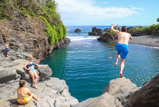 Hana, Hawaii - February 2020: A young man leaps from a cliff ledge into Maui's Waioka Pond, aka the Venus Pool, in front of several other visitors.