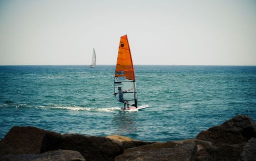 Two sailboats on a sunny day, gliding across a body of water near a dock and rocky shore