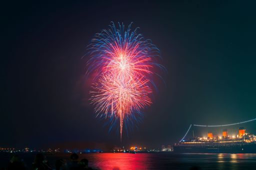 A vibrant display of fireworks illuminates the night sky over a peaceful body of water, with the Queen Mary in the foreground