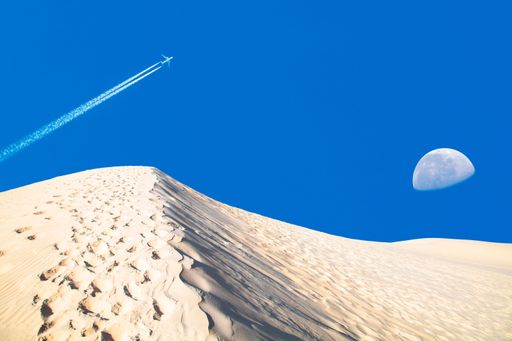 The White sand dunes and blue sky with half moon and plane with contrail.