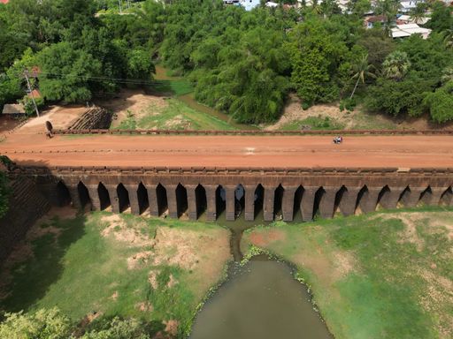 Aerial view of a rustic unpaved road meandering through verdant trees and crossing a picturesque bridge in a serene forest setting