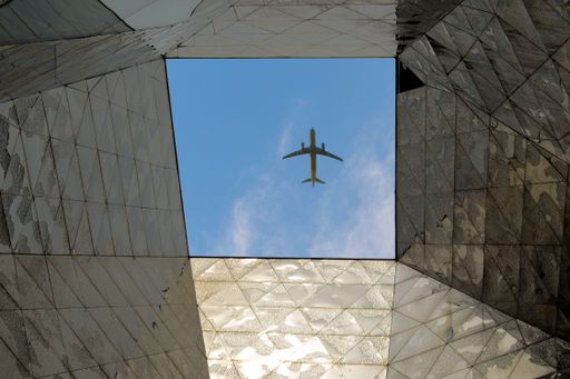 An airplane flying in the blue sky of a sunny day seen from the ground through the hole in the roof of a building