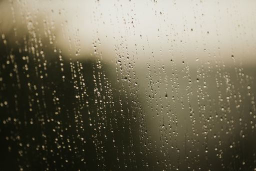 A blurry shot of a glass window covered with raindrops and a dimly lit background.