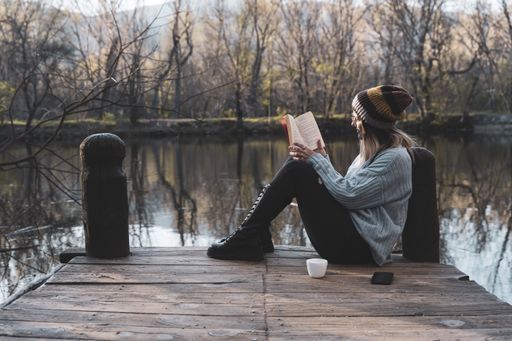 a young woman sits on a jetty above the lake, she is reading a book