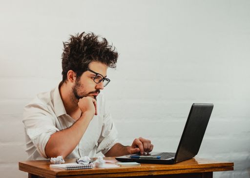 A young Caucasian male staring at the laptop, bored and disheveled during work, resting head on hand