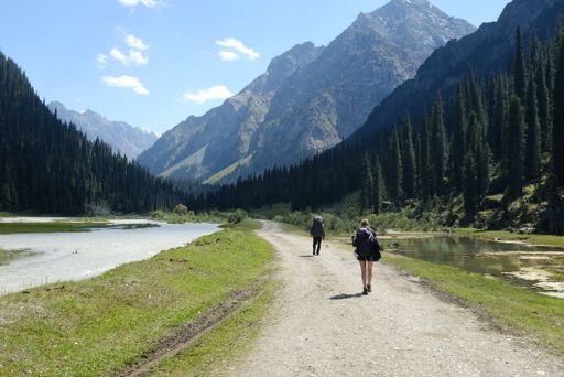 The two people walking along a winding dirt road surrounded by majestic mountains.