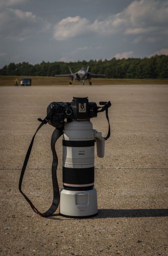 A mirrorless digital camera with a super zoom lens standing up at an airport with a fighter jet in the background