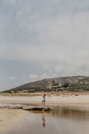 A vertical shot of a woman standing on the shore with mountains on the background