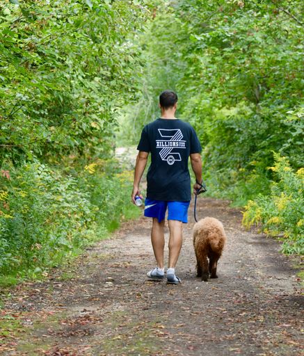 A man and his loyal canine companion are enjoying a leisurely stroll through a peaceful wooded area, surrounded by lush foliage