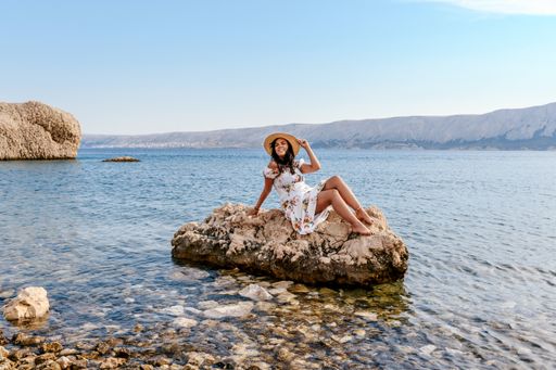 Woman in elegant white summer dress sitting on rock on beautiful Beritnica beach on Pag island in Croatia.