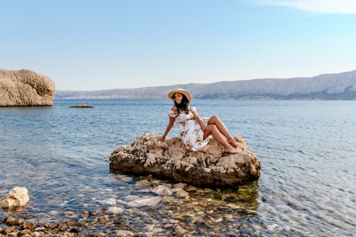 Woman in elegant white summer dress sitting on rock on beautiful Beritnica beach on Pag island in Croatia.