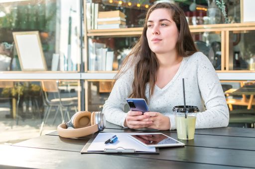 A thoughtful young woman using her smartphone while drinking juice and working in the cafe
