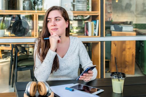 A thoughtful young woman using her smartphone while drinking juice and working in the caf