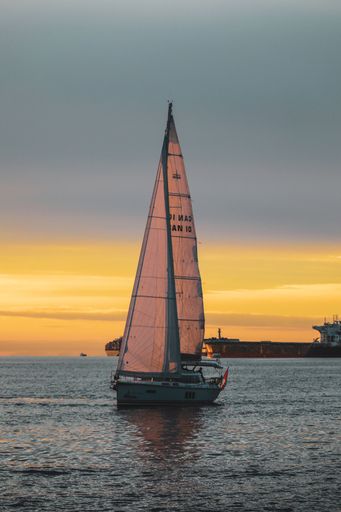 A vertical shot of a sailing boat in the lake during sunset with a ship in the background