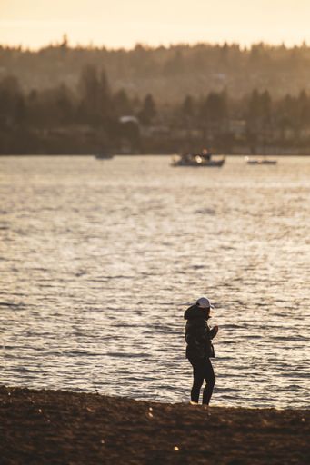 A vertical shot of the woman near the shore with boats and mountains in the background during sunset
