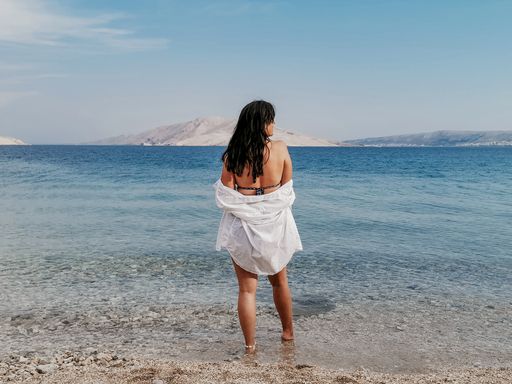 Rear view of stylish young woman wearing only white shirt on beach and looking at view on Beritnica beach on Pag island in Croatia