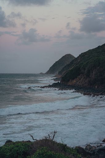 A vertical shot of a wavy ocean beach with cliffs and hills at sunset