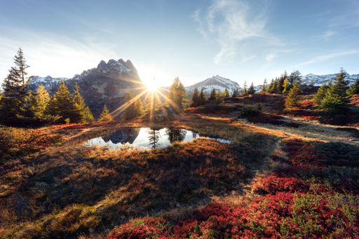 Landscape autumnal mountain view of a small pond surrounded by blueberry bushes with a sunburst between the peaks in Emosson Wallis, Switzerland