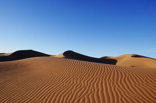 A high-angle shot of a vast desert landscape featuring rolling sand dunes, a bright blue sky, and a dramatic horizon