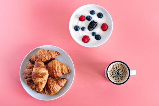 A top view of a pink breakfast table with coffee, croissants, and a bowl of berries with yogurt