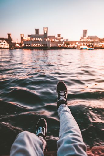 A vertical shot of a man sitting on a border of a lake with blurred city lights on a background