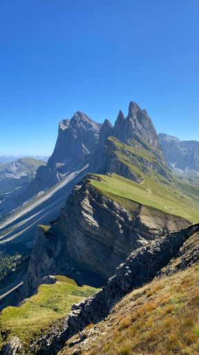 An expansive view of a mountain range on a sunny day, with lush green grass and rocky terrain: Secada, Italy