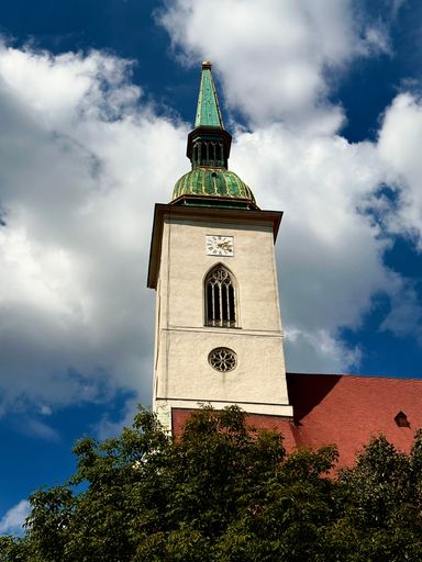 A picturesque view of a traditional church tower surrounded by lush green trees set against a clear blue sky with white fluffy clouds