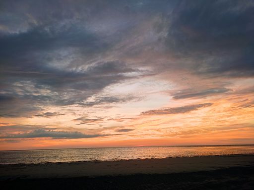 A spectacular evening beach scene featuring a vibrant orange and red sky, illuminated by the setting sun, with dark clouds looming in the background