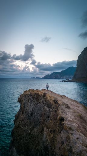 A young man stands atop a rocky cliff overlooking the expansive ocean