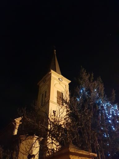 A majestic lit church tower illuminated against the dark night sky, standing tall with a decorated Christmas tree at the base of the tower