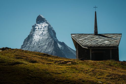 An elevated view of a traditional church situated atop a grassy hill with a majestic mountain range in the background