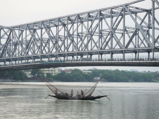 Two men in a traditional boat row across the Hoogly river with the Howrah bridge in the background for fish