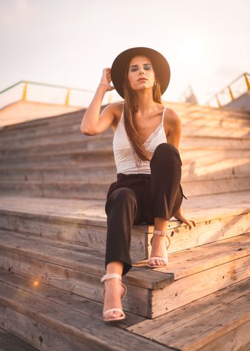 A beautiful brunette female in a black hat sitting on stairs and posing during the sunset