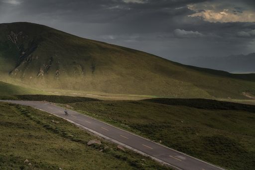 An aerial view of a road amid green fields in the background of hills under a cloudy sky