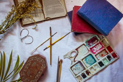 A white bedsheet with art supplies and leather-bound journals on it around leaves and a jewelry box