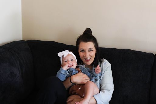 A young mother cradles her infant child in her arms while seated on a black sofa against a solid wall background