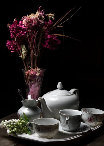 A vertical shot of ceramic teacups and kettle with dried flowers in a vase on a black background