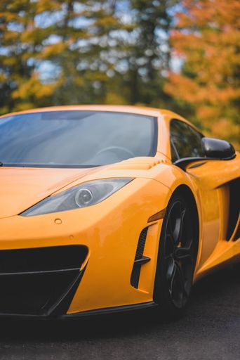 An orange luxury sports car driving on a scenic rural road surrounded by lush green trees