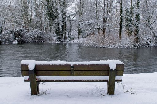 A wide angle shot of a wooden bench in front of the water in a park covered in snow