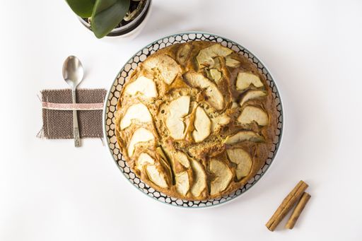 A top view of a delicious apple pie on a plate on a white surface with cinnamon sticks and a spoon on it